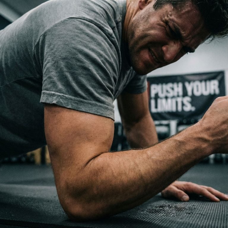 팔굽혀펴기를 하는 근육 팽팽한 등과 팔에 땀방울이 맺힌 채, 고통스럽지만 의지가 담긴 얼굴을 가까이서 담은 모습이다.